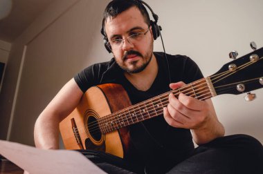 Sakallı, kulaklıklı ve gözlüklü, siyah giyinmiş ve evdeki oturma odasında yere oturmuş gitar çalıyor, nota okuyor..