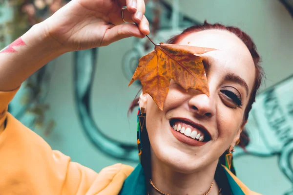 Güzel, genç, Latin, Arjantinli bir kadın gülüyor bir gözünü kuru bir sonbahar yaprağıyla kaplıyor, kameraya bakıyor mutlu ve neşeli..