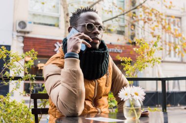 Afrikalı yetişkin bir adam dışarıda bir kafede oturuyor, telefonla konuşuyor, fotokopi odası, teknoloji konsepti..