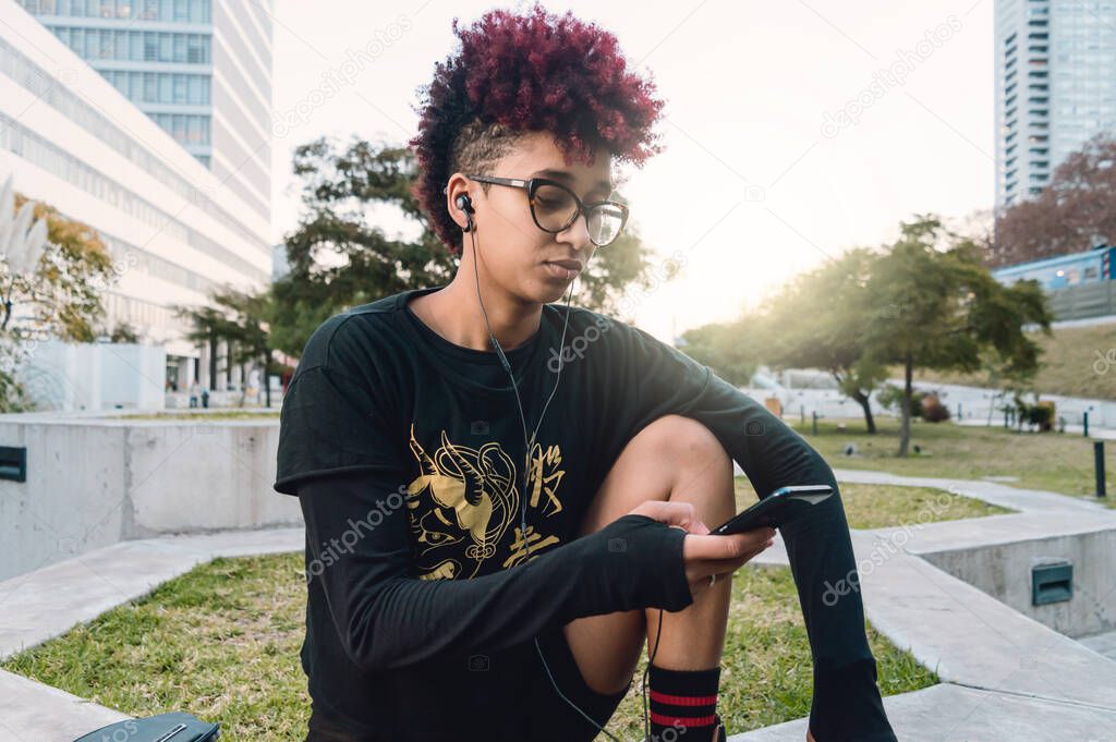 young latina, hispanic colombian brunette woman with afro and glasses, sitting outdoors at sunset with headphones looking for music on her phone