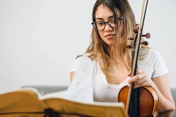 young latin caucasian violinist woman, with glasses ethnic venezuelan long hair, at home studying sheet music sitting on sofa with violin in hand, copy space.