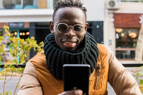 happy african man alone outdoors, smile and check notifications and messages from social networks and mail on his phone while waiting for breakfast, sitting outside cafeteria, front view.