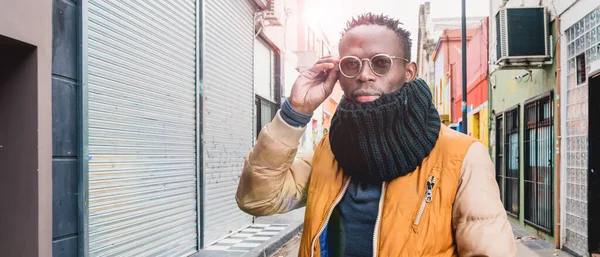 banner of young african man outdoors, standing in the middle of the street, dressed in winter clothes, looking at the camera touching his glasses with one hand, urban environment with copy space