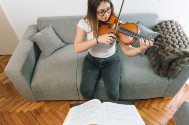 Ön manzara, gözlüklü, keman çalan, evdeki kanepede oturan genç Latin, beyaz Venezuelalı kadın. Klasik müzik çalışmak ve çalışmak..