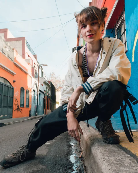 young latin trans woman, posing looking at the camera crouched on the edge of the sidewalk with copy space on the left, vertical image, urban fashion concept.