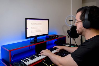 caucasian latin young man, with black clothes, headphones, beard and glasses, studying and practicing electric piano at home by online classes, sitting in his home studio. clipart