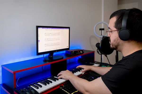 caucasian latin young man, with black clothes, headphones, beard and glasses, studying and practicing electric piano at home by online classes, sitting in his home studio.