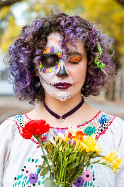 vertical portrait of young woman with La Calavera Catrina makeup and closed eyes, holding a bouquet of flowers standing in the cemetery.