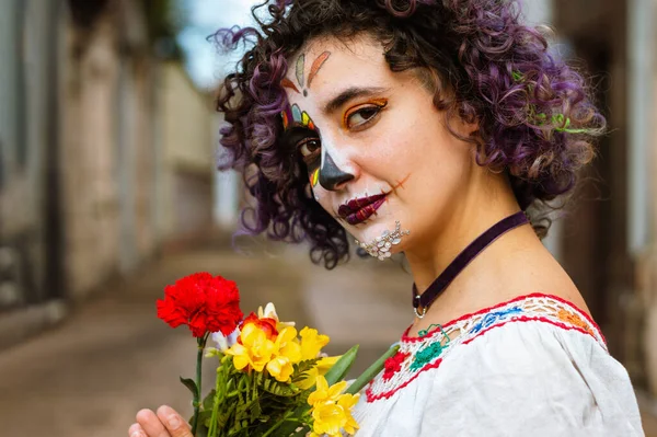 portrait of young latin caucasian argentinian woman, holding flowers with her hands standing outdoors in the cemetery with La Calavera Catrina makeup, looking at the camera in profile.