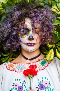 vertical portrait of young latin woman with closed eyes, purple curly hair and La Calavera Catrina makeup, on a background of natural leaves outdoors