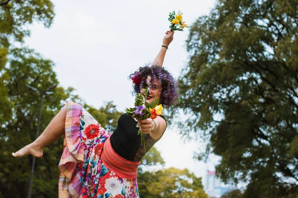 young latin caucasian woman with flowers in her hands dancing in the park with colorful skirt and La Calavera Catrina makeup, smiling and looking at the camera with one leg raised and copy space.