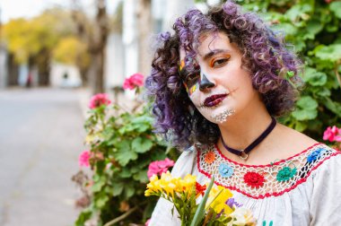 La Calavera Catrina makyajlı ve folklorik elbiseli Latin Arjantinli genç bir kadın kameraya dalgın bir suratla bakıyor ve elinde çiçekler tutuyor.