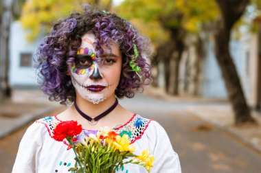 Elinde çiçekleri olan ve sanatsal makyajı La Calavera Catrina 'yı temsil eden, açık havada fotokopi aletiyle kameraya bakan ciddi bir genç kadının portresi..