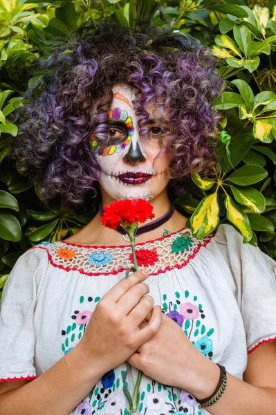 照片上年轻的拉丁女子站在室外 手里拿着一朵花 她的脸被漆成卡特里娜 卡特里娜 背景是天然绿叶 — 图库照片