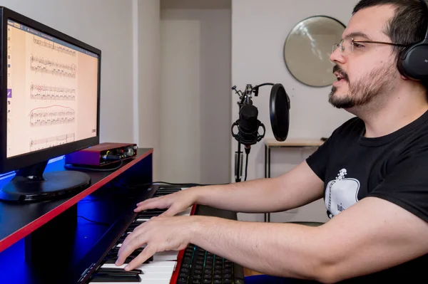 Sakallı, gözlüklü, kulaklıklı, piyano çalıp şarkı söyleyen, evdeki bilgisayarla dijital müzik okuyan genç beyaz bir adam.