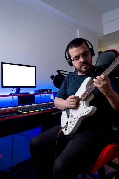 bearded male rock music producer in glasses and headphones, recording the electric guitar sitting in his home music studio.