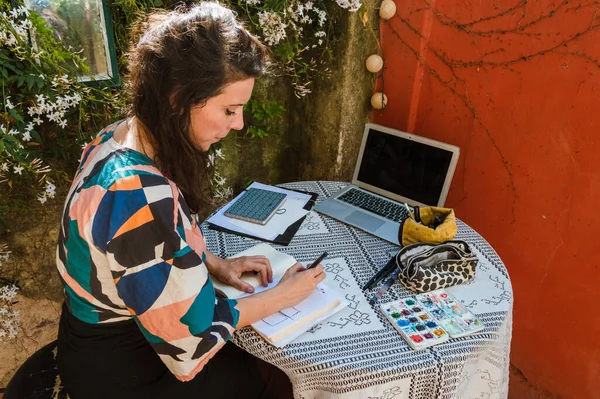 Caucasian french adult woman, designer working at home with the computer, making sketches on paper, with watercolor and drawing implements on the table.