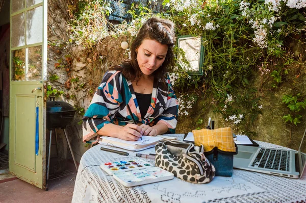 Masaya oturan beyaz bir kadın evinin terasında defter ve suluboya boyayla arka planda zambaklar olan bir duvar çiziyor.