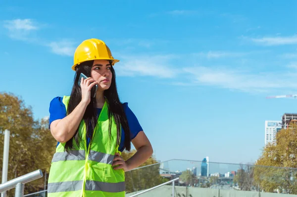 young latin venezuelan engineer woman with yellow helmet, vest and blue clothes, standing outdoors with a hand on her waist and on a phone call waiting for a response from her boss to start work.