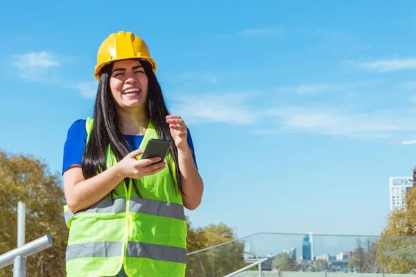young latin venezuelan engineer woman, wearing yellow helmet and vest standing outdoors laughing out loud because of a joke texted to her phone, technology and communication concept, copy space.
