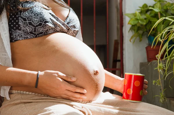 お腹に手を触れながら家に座っている若いラテン系の妊婦の近くで — ストック写真