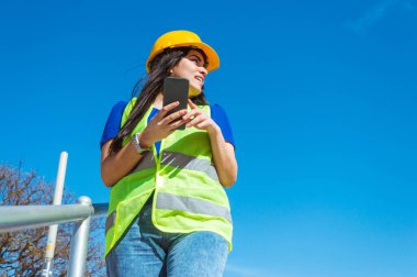 Güvenlik kaskı takan genç, beyaz bir kadın dışarıda durup telefonunu tutarken biriyle tartışıyor. Boşluğu kopyala.