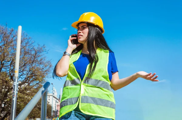 young caucasian venezuelan woman, standing outdoors wearing yellow safety helmet, blue clothes and vest, is annoyed arguing on the phone with her boss.