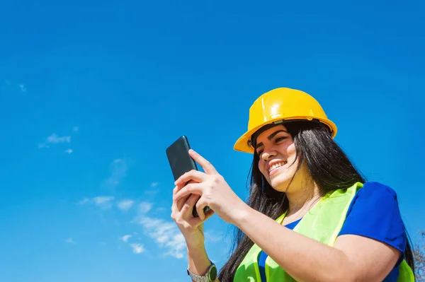 young latin, caucasian, venezuelan woman worker with yellow safety helmet outdoors happy smiling checking her phone, reading notifications, with blue sky in the background and copy space.