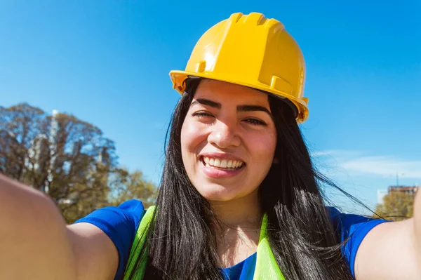 portrait of a young caucasian venezuelan worker woman, wearing blue clothes, yellow safety helmet and vest is outdoors smiling taking a selfie with her cell phone