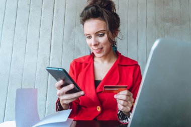 Kırmızılı, dizüstü bilgisayarı ve telefonuyla oturmuş, kredi kartıyla internet ödemeleri yapan, ön planda bilgisayarı olan genç serbest çalışan bir kadının ön görüntüsü..