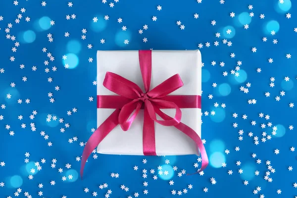 Подарочная коробка завернутая в белую бумагу с фиолетовым бантом на праздничном голубом фоне со многими снежинками — стоковое фото