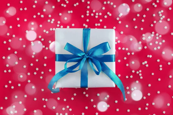 Подарочная коробка завернутая в белую бумагу с голубым бантом на праздничном малиновом фоне со многими снежинками — стоковое фото