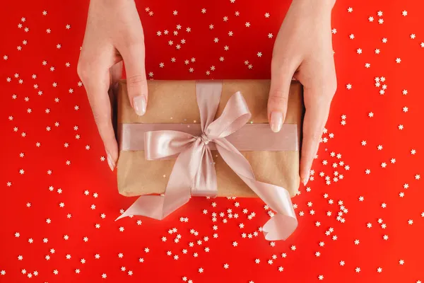 Mooie vrouwelijke handen houden geschenkdoos verpakt in kraftpapier met roze lint op feestelijke achtergrond — Stockfoto