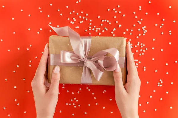 Mooie vrouwelijke handen houden geschenkdoos verpakt in kraftpapier met roze lint op feestelijke achtergrond — Stockfoto