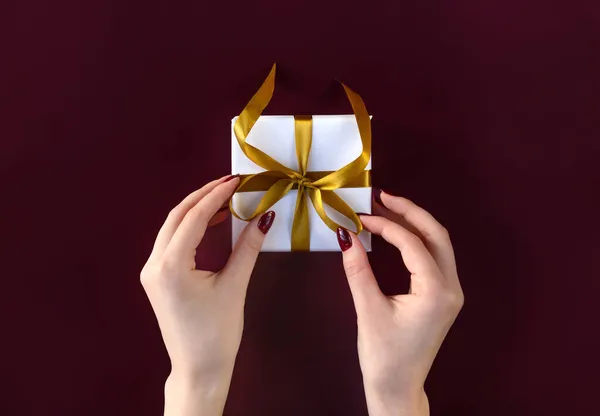 Jolies mains féminines tenant boîte cadeau enveloppée dans du papier blanc avec ruban doré sur fond de fête — Photo