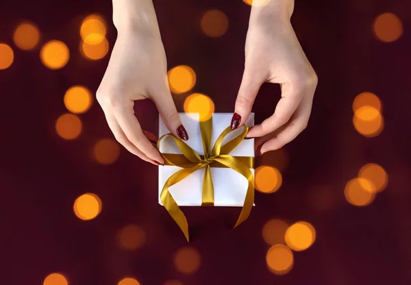 Jolies mains féminines tenant boîte cadeau enveloppée dans du papier blanc avec ruban doré sur fond de fête — Photo