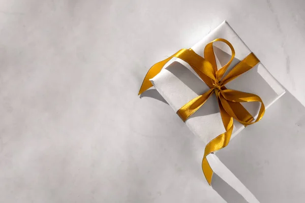 Coffret cadeau enveloppé dans du papier blanc avec un arc doré sur fond de lumière festive — Photo