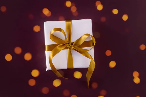 Подарочная коробка, завернутая в белую бумагу с золотым бантом на праздничном фиолетовом фоне — стоковое фото