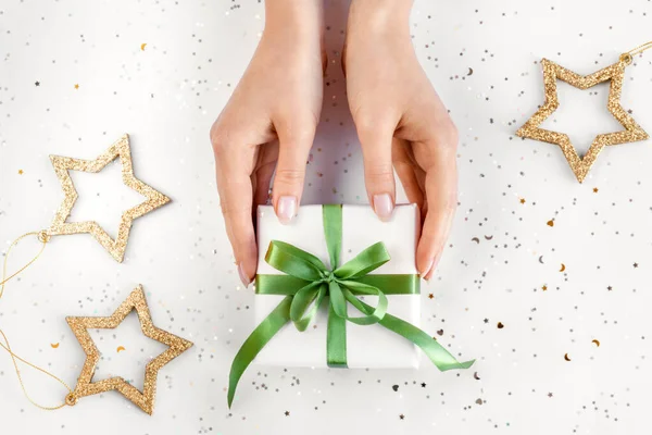 Vrouwelijke handen met natuurlijke roze manicure houden een geschenk in wit papier met groen lint. Kerstcompositie. Vlakke plaat — Stockfoto