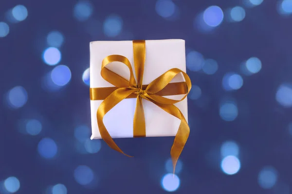 Coffret cadeau enveloppé dans du papier blanc avec un noeud doré sur fond bleu violet festif — Photo