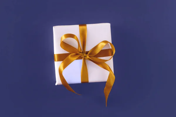 Подарочная коробка, завернутая в белую бумагу с золотым бантом на праздничном фиолетовом синем фоне — стоковое фото