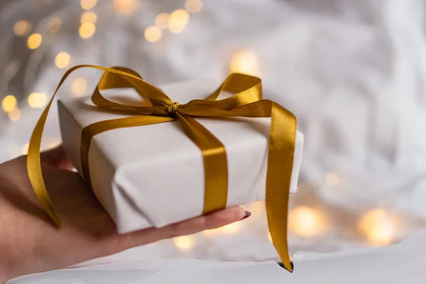Mano con caja de regalo envuelta en papel blanco con un lazo dorado sobre fondo de luz festiva con luces bokeh —  Fotos de Stock