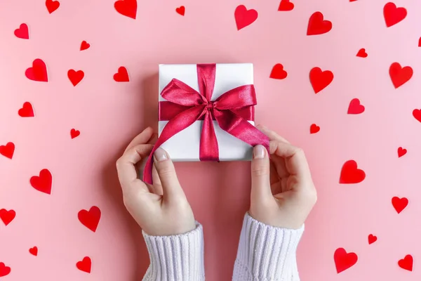 Female Hands holding a gift in white wrapping paper on pink background with red hearts — Stock Photo, Image