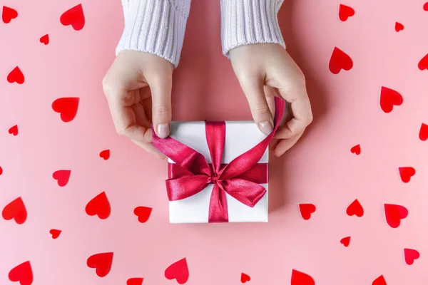 Mãos femininas segurando um presente em papel de embrulho branco no fundo rosa com corações vermelhos — Fotografia de Stock
