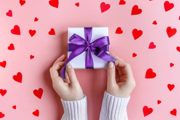 Female Hands holding a gift in white wrapping paper on pink background with red hearts — Stock Photo, Image