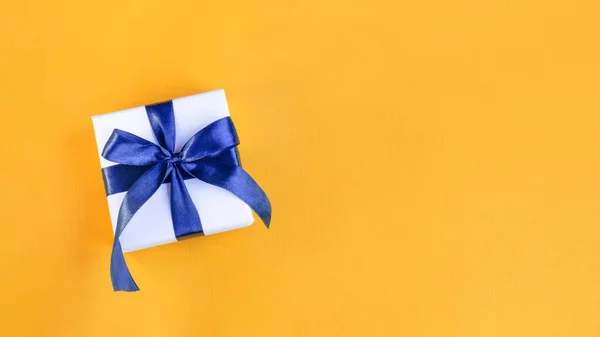 Bannière d'un cadeau enveloppé dans du papier blanc avec un arc bleu en satin sur fond orange jaune festive — Photo