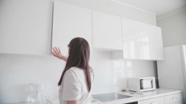 Genç bir kadın kapıyı açar ve dolaptan cam bardakları çıkarır. Beyaz tişörtlü ve kot pantolonlu, kollarını üst rafa uzatıyor. Siyah saçlı ve gözlüklü kız mutfakta beyaz mutfak takımıyla. 