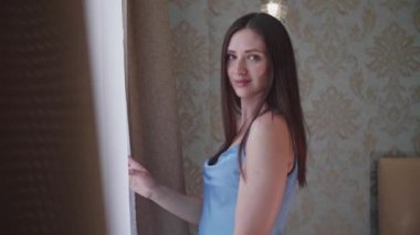 Yatak odasındaki pencerenin yanında duran genç kadın gülümsüyor ve güneşin doğuşunu seyrediyor. O, narin mavi renkli gömlek giydi. Beyaz tüllü pencere. Sabah, şafak, dinlenme, yeni bir günün başlangıcı