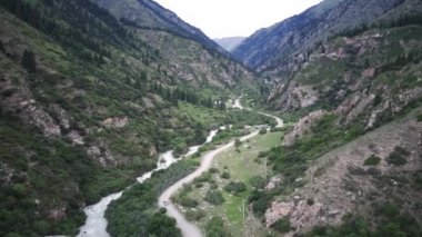 Bir araba, yeşil ormanı, vahşi nehri ve yurdu geçen bir helikopter tarafından takip ediliyor.