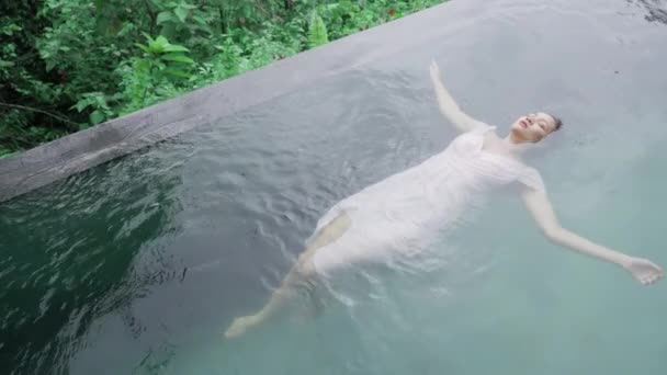 Vista top shot de uma menina de raça mista em um vestido de noite rosa suave que está nadando de costas em uma piscina com água verde esmeralda em tempo nublado. Uma menina asiática está deitada de costas em uma piscina. — Vídeo de Stock